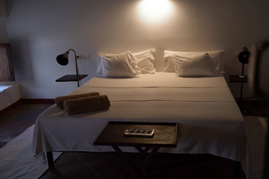 Acogedor dormitorio con una cama grande, sábanas blancas, mesitas de noche y lámparas. Dos toallas enrolladas descansan sobre la cama.