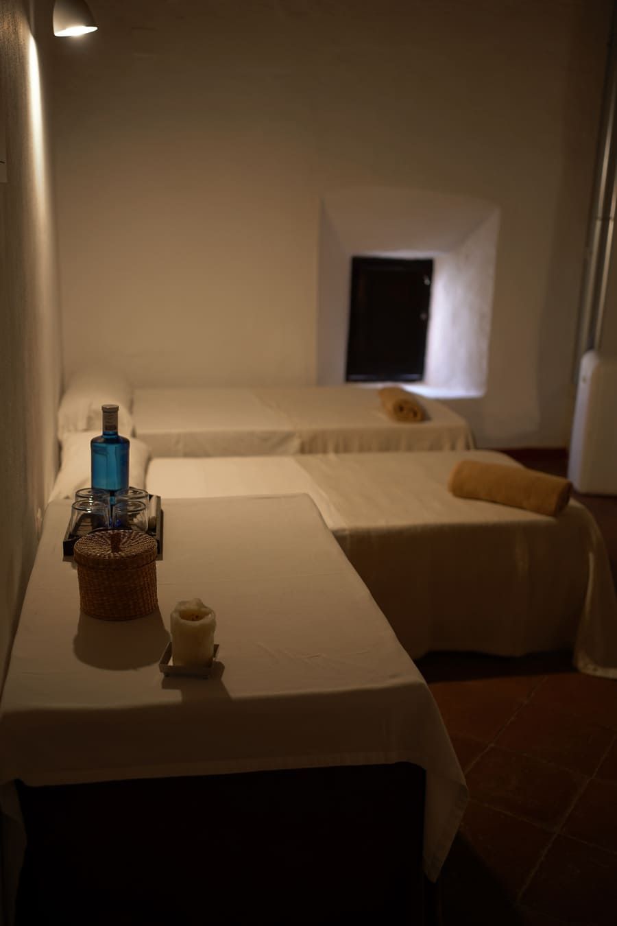 Sala de masajes con dos camas, sábanas blancas y velas, bañada por una suave luz.