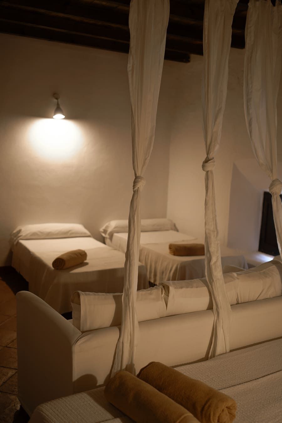 Habitación de paredes blancas con varias camas, algunas con tela blanca. Toallas enrolladas sobre las camas.