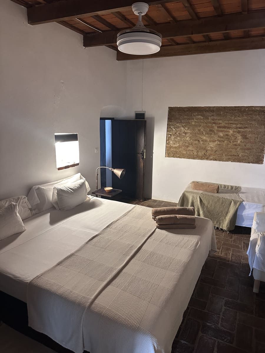 Dormitorio con cama grande blanca, armario oscuro y asientos; estilo rústico.