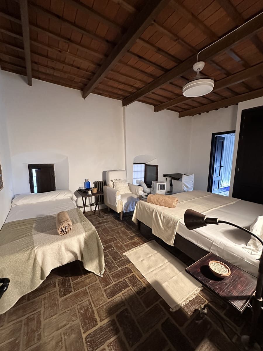 Dormitorio con piso de ladrillo, paredes blancas, techo de madera y dos camas con mantas beige.