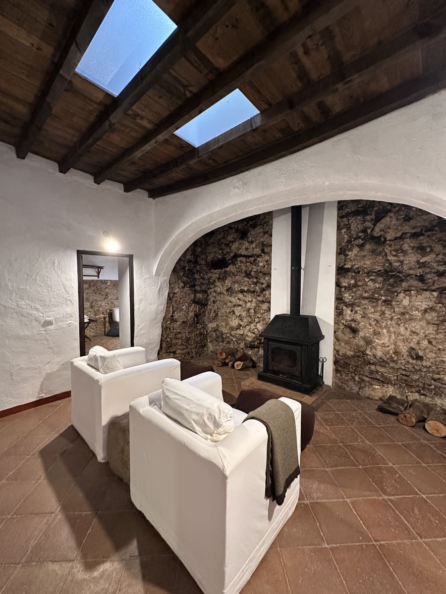 Acogedora sala de estar con sillones blancos, chimenea, pared de piedra vista y tragaluces en un entorno rústico.