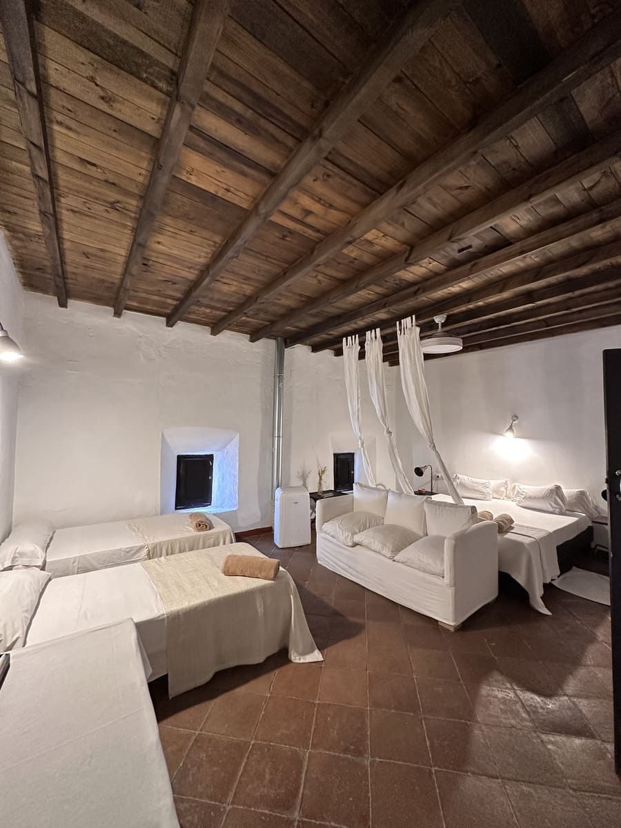 Dormitorio con paredes blancas, techo de madera, piso de baldosas rojas, varias camas y un sofá blanco.