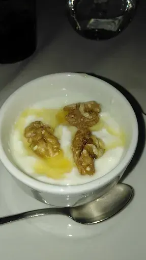 Tazón blanco con yogur, nueces y miel, sobre un plato blanco con una cuchara.