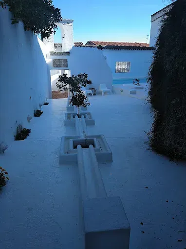 Patio blanco con pequeños árboles y piscina, bajo un cielo azul.