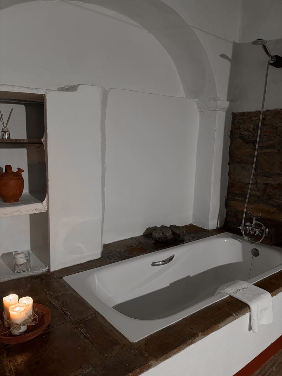 Baño con bañera blanca, estante de madera, velas y pared de piedra.