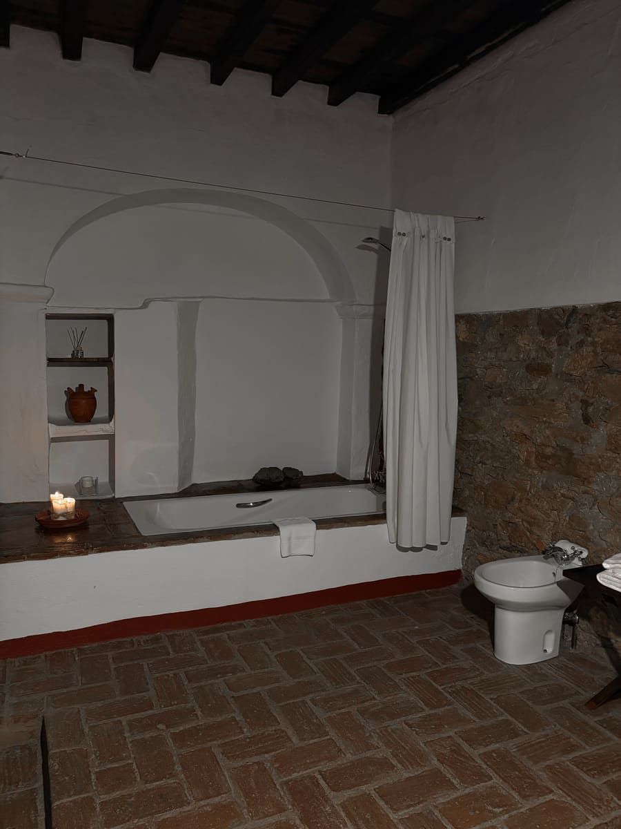Baño con bañera incorporada, pared de piedra, suelo de ladrillo y vela encendida.