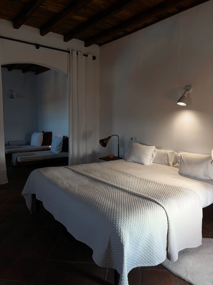 Acogedor dormitorio con una gran cama blanca, una zona de estar al fondo y una iluminación cálida.