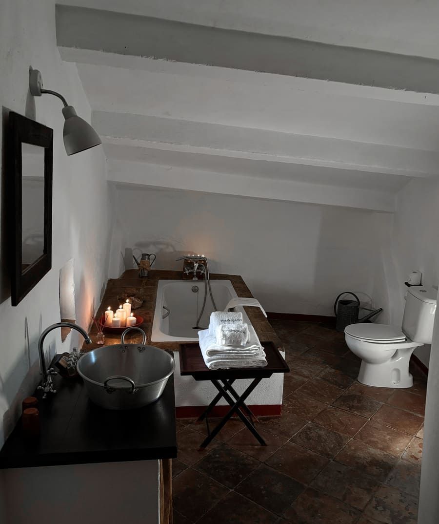 Baño rústico con bañera, lavabo e inodoro bajo un techo inclinado. Velas encendidas.