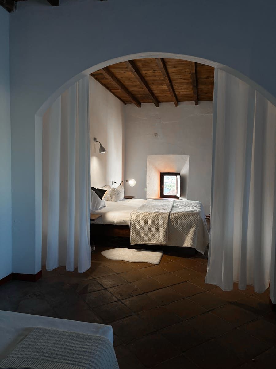 Dormitorio con paredes blancas, cortinas de arco, cama y pequeña ventana.