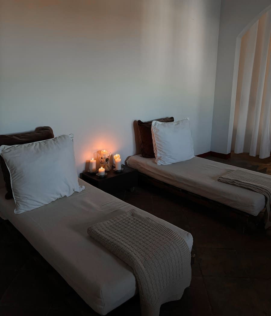 Dos camas con sábanas y almohadas blancas en una habitación poco iluminada, con velas en una mesita de noche.