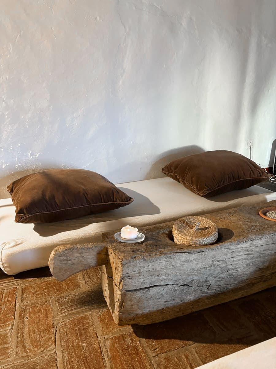 Banco bajo de madera con cojines marrones contra una pared blanca, entorno rústico.