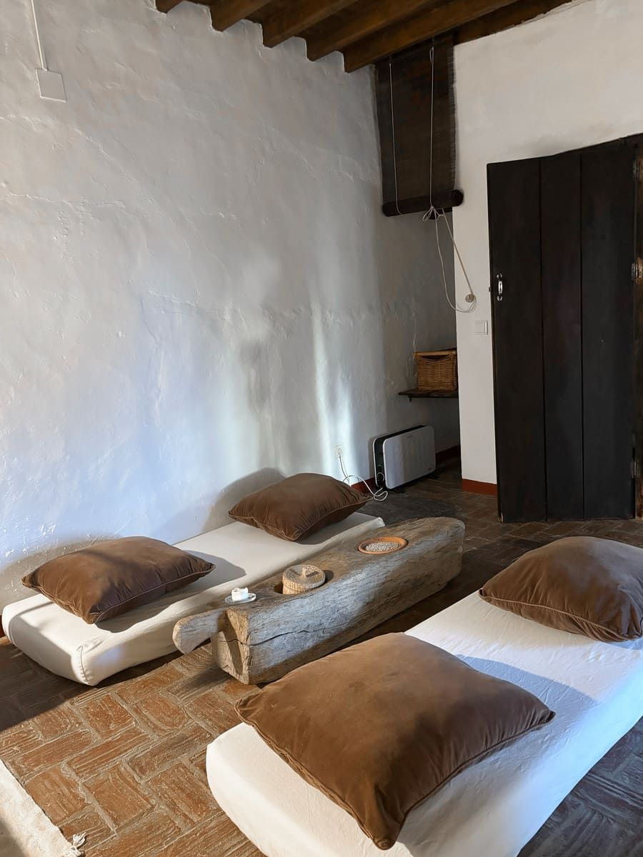 Habitación de paredes blancas con dos camas, almohadas marrones, una mesa con viga de madera y una puerta de madera oscura.