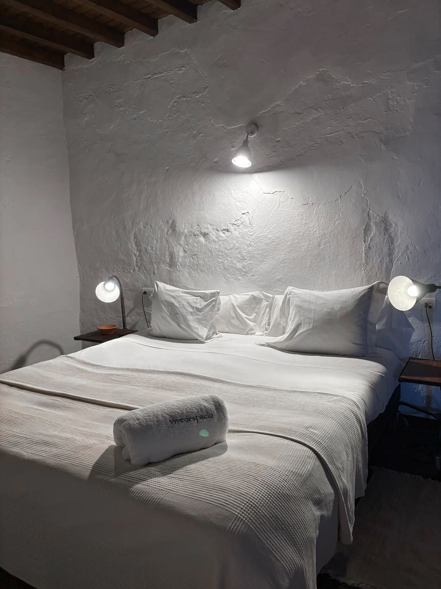Dormitorio de paredes blancas con cama, almohadas, manta y toalla enrollada. Dos lámparas de noche y luz de techo.
