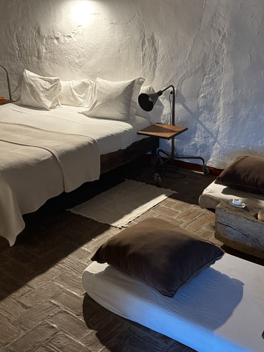 Dormitorio con cama, mesita y plataformas acolchadas. Paredes blancas, ropa de cama color crema y suelo marrón.