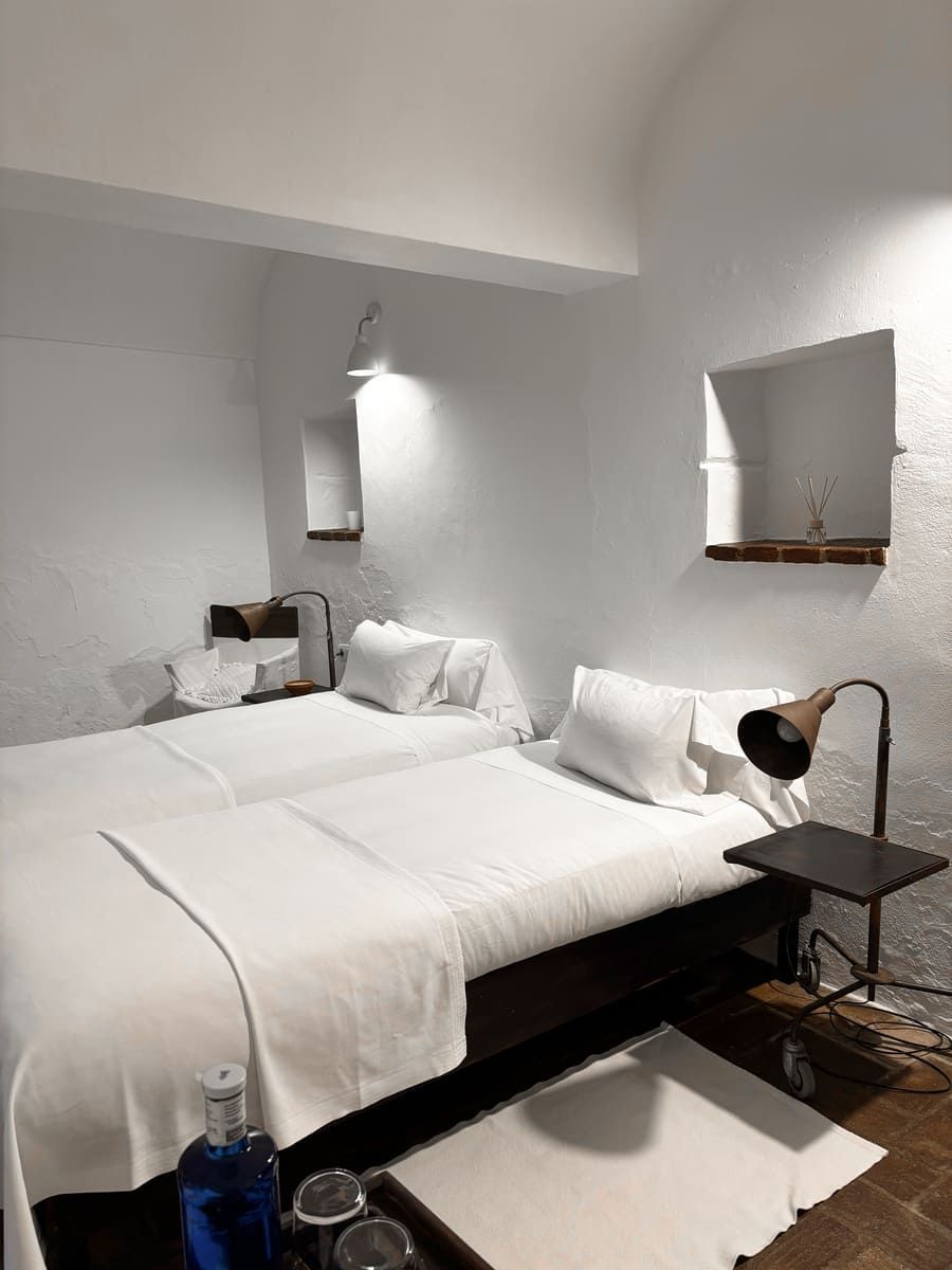 Dormitorio de paredes blancas con dos camas, mesita de noche y una pequeña ventana. Diseño minimalista.