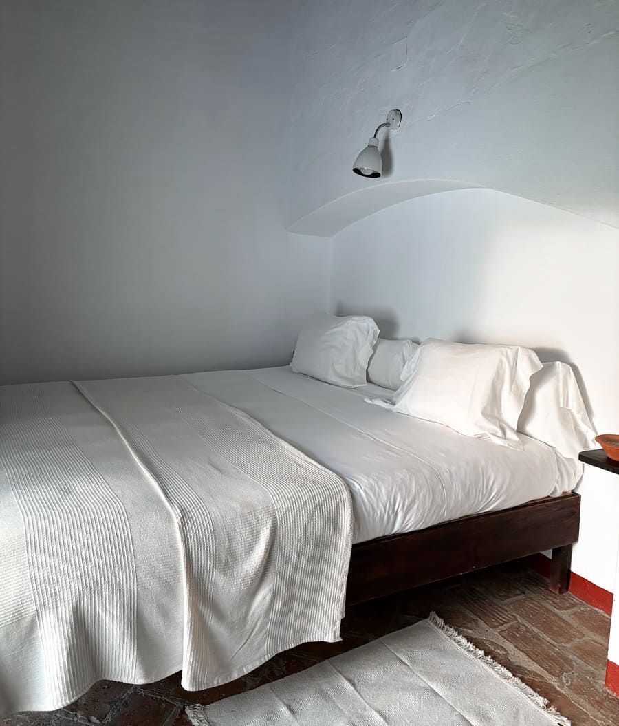 Cama blanca con ropa de cama blanca en una habitación pequeña; paredes blancas; la cama está al lado de una superficie roja; lámpara de techo.