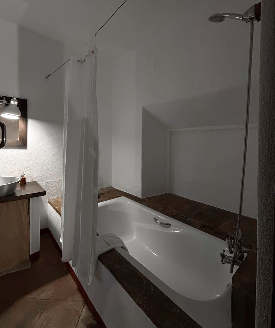 Baño con bañera blanca, ducha y cortina. Molduras de madera alrededor de la bañera, paredes blancas y azulejos de terracota.