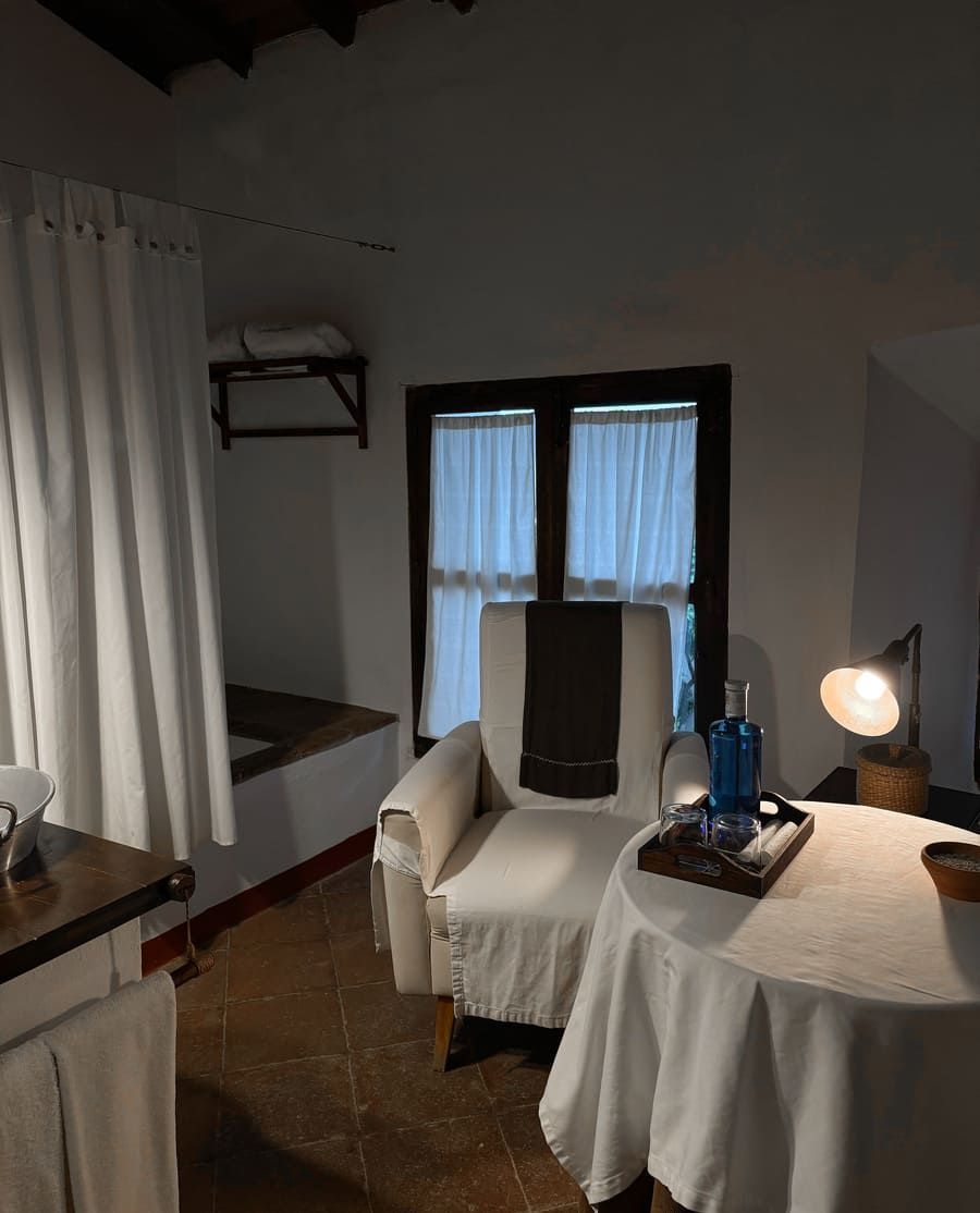 Acogedora habitación con un sillón blanco, una mesa pequeña y una ventana con cortinas transparentes.