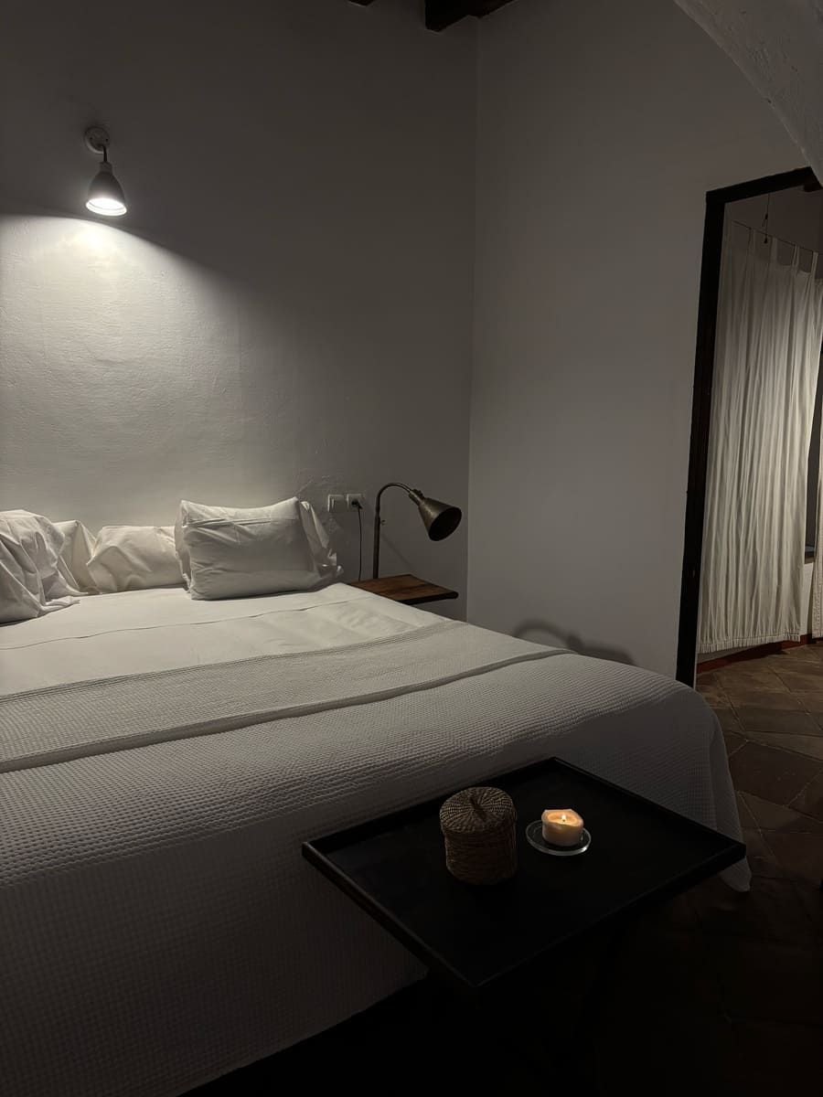 Dormitorio con ropa de cama blanca, lámpara de noche y bandeja con vela. Iluminación tenue.