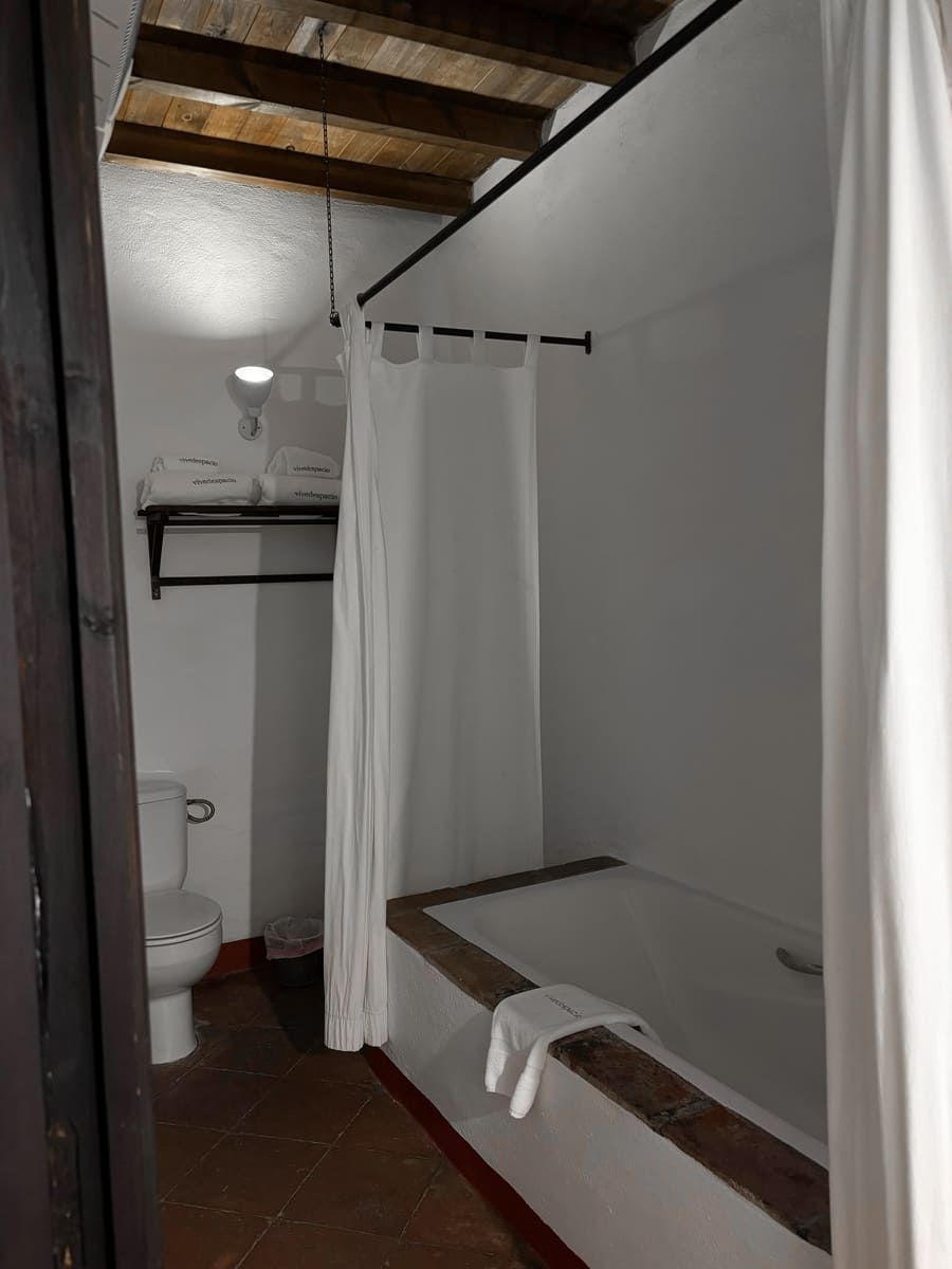 Baño con bañera, inodoro, toalleros en perchero, paredes blancas y vigas de madera a la vista en el techo.