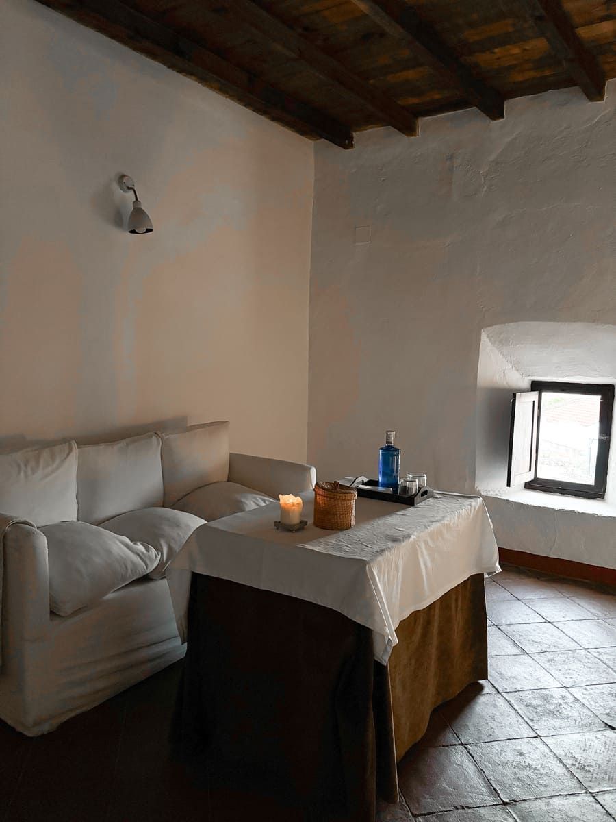 Acogedora sala de spa: sofá blanco, mesita con vela y botella de agua, ventana. Suelo marrón oscuro.