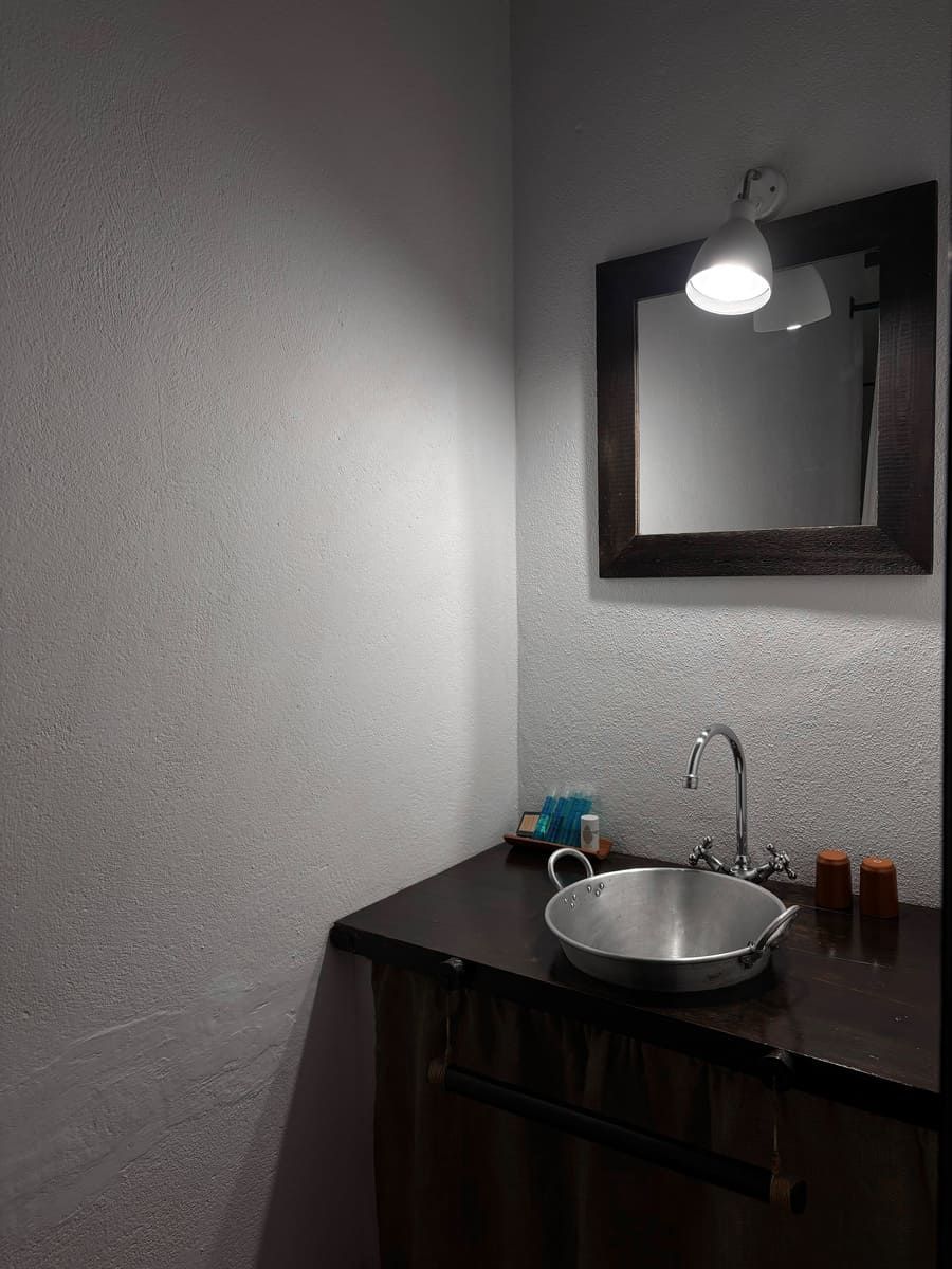 Baño de esquina con tocador de madera oscura, lavabo metálico, espejo y luz de techo.