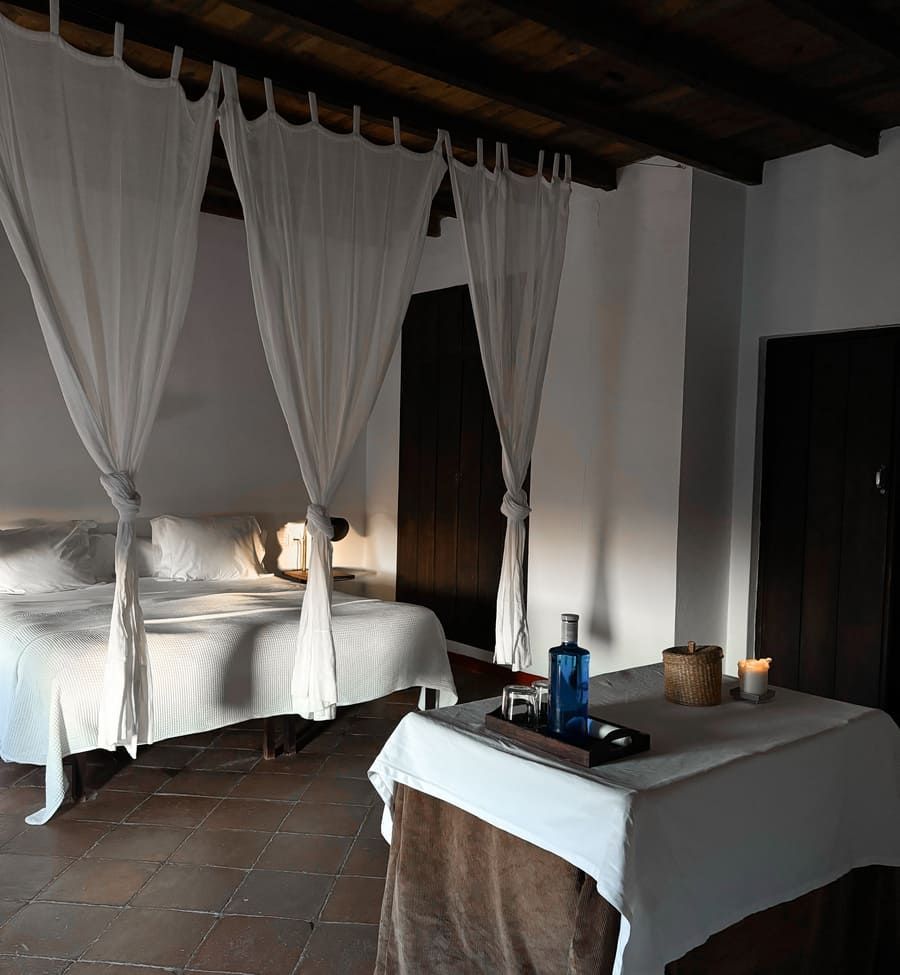 Dormitorio con cama con dosel blanco, suelo de terracota, mesa puesta con botella de agua y vasos.