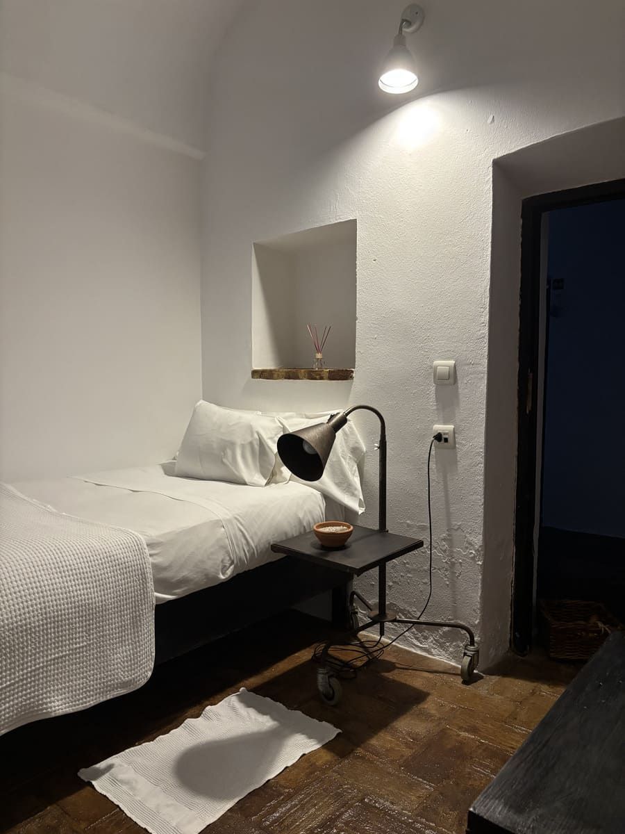 Una pequeña habitación blanca con cama, mesita de noche y puerta. Lámpara encendida, sombras proyectadas en el suelo.