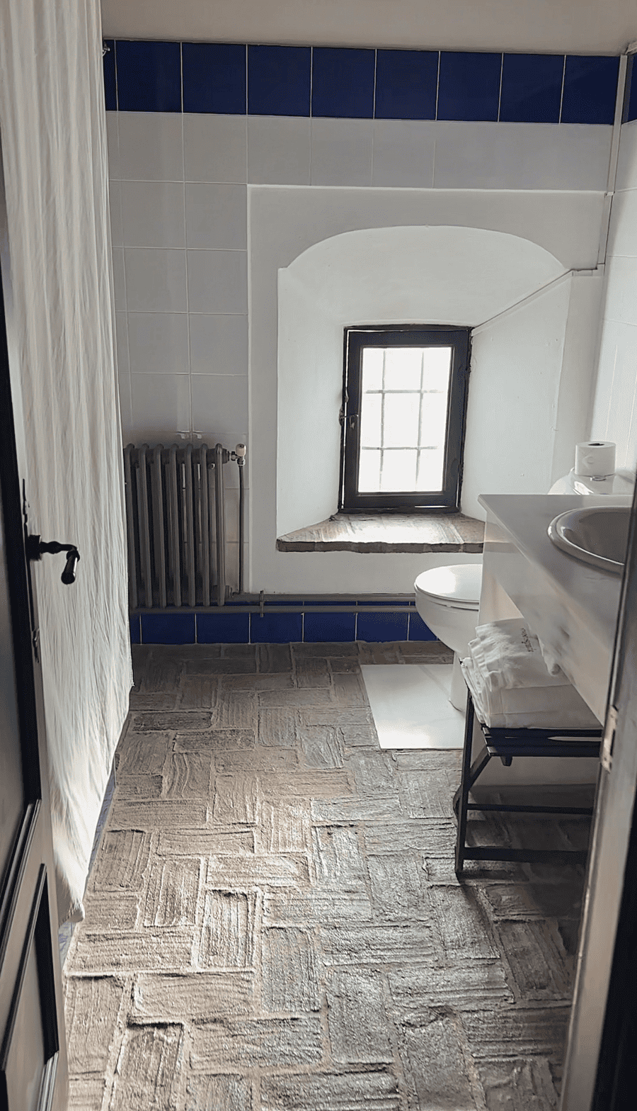 Baño con suelo de baldosas, ventana, inodoro, radiador y paredes blancas con detalles en azul.