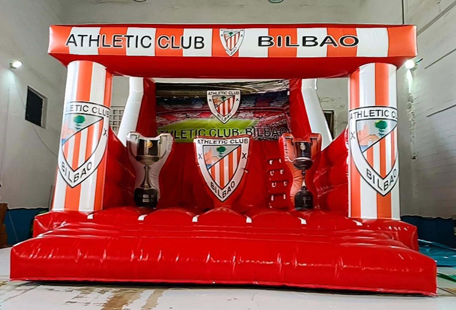 Un escenario inflable del Athletic Club Bilbao con temática de fútbol, con trofeos, el logo del equipo y el fondo del estadio. Predominantemente rojo y blanco.