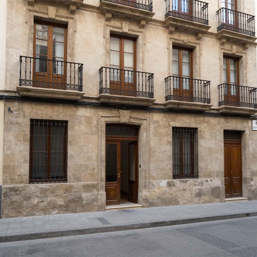 Fachada de piedra de un edificio de apartamentos, con balcones, ventanas con rejas y dos puertas de madera, en una calle tranquila.