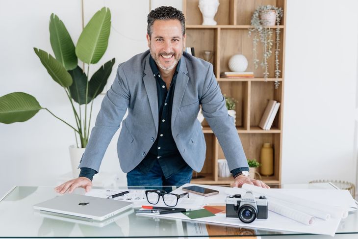 Hombre sonriendo detrás de un escritorio con computadora portátil, papeles, cámara y gafas en una oficina luminosa.