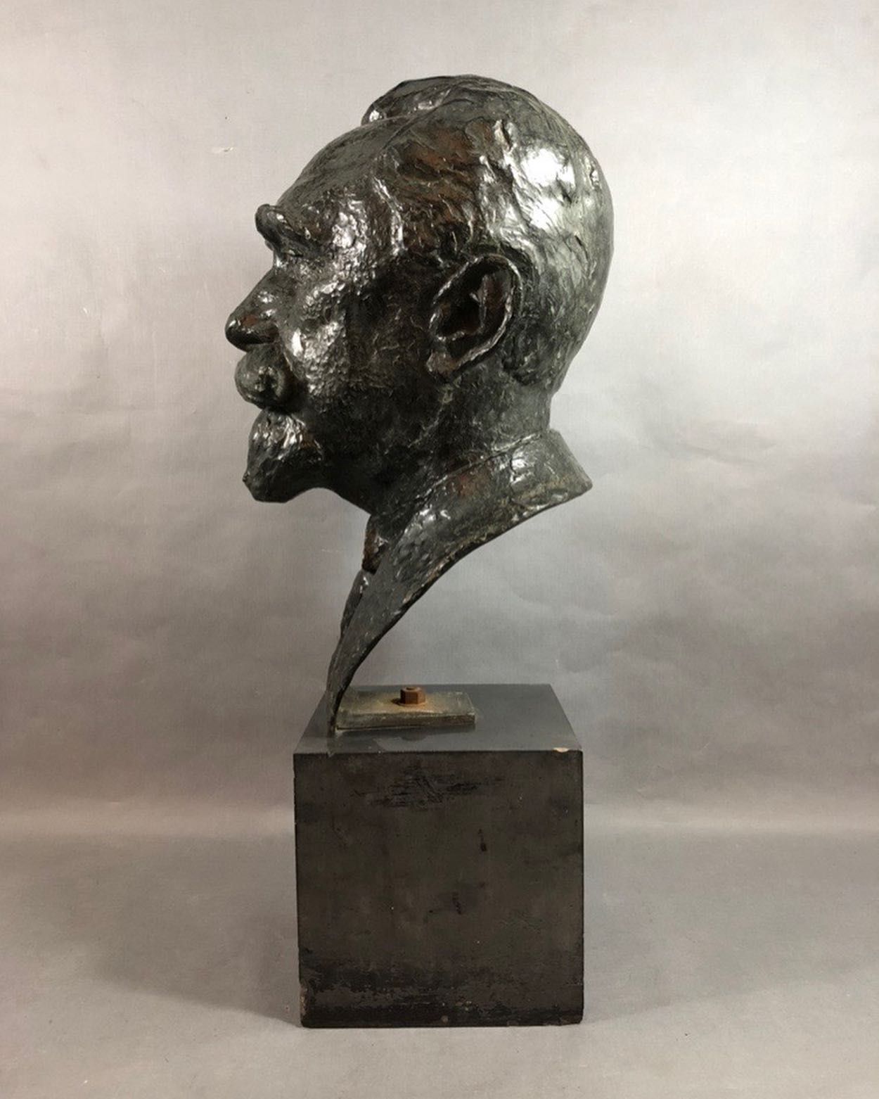 Busto de bronze de um homem com bigode, montado sobre um bloco quadrado escuro.