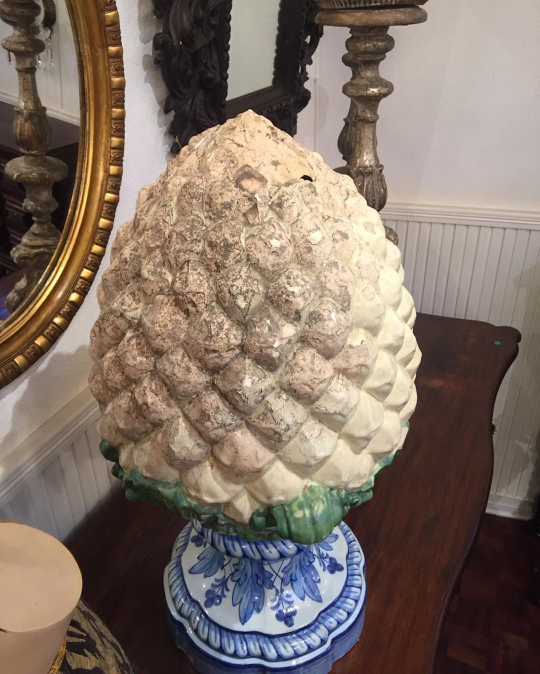 Escultura grande e decorativa em forma de pinha com escamas bege-claras, sobre uma base de cerâmica azul e branca.