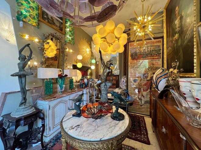 Interior de loja de antiguidades com móveis ornamentados, esculturas e luminárias; diversas cores.