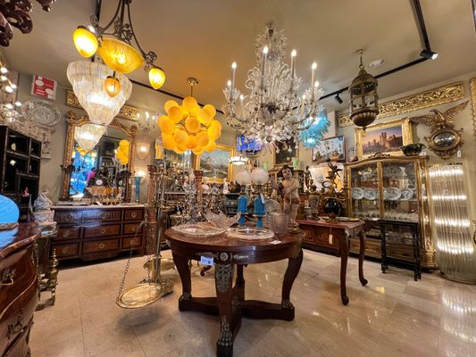 Interior de uma loja de antiguidades repleta de lustres e móveis elaborados, com uma mesa redonda no centro.