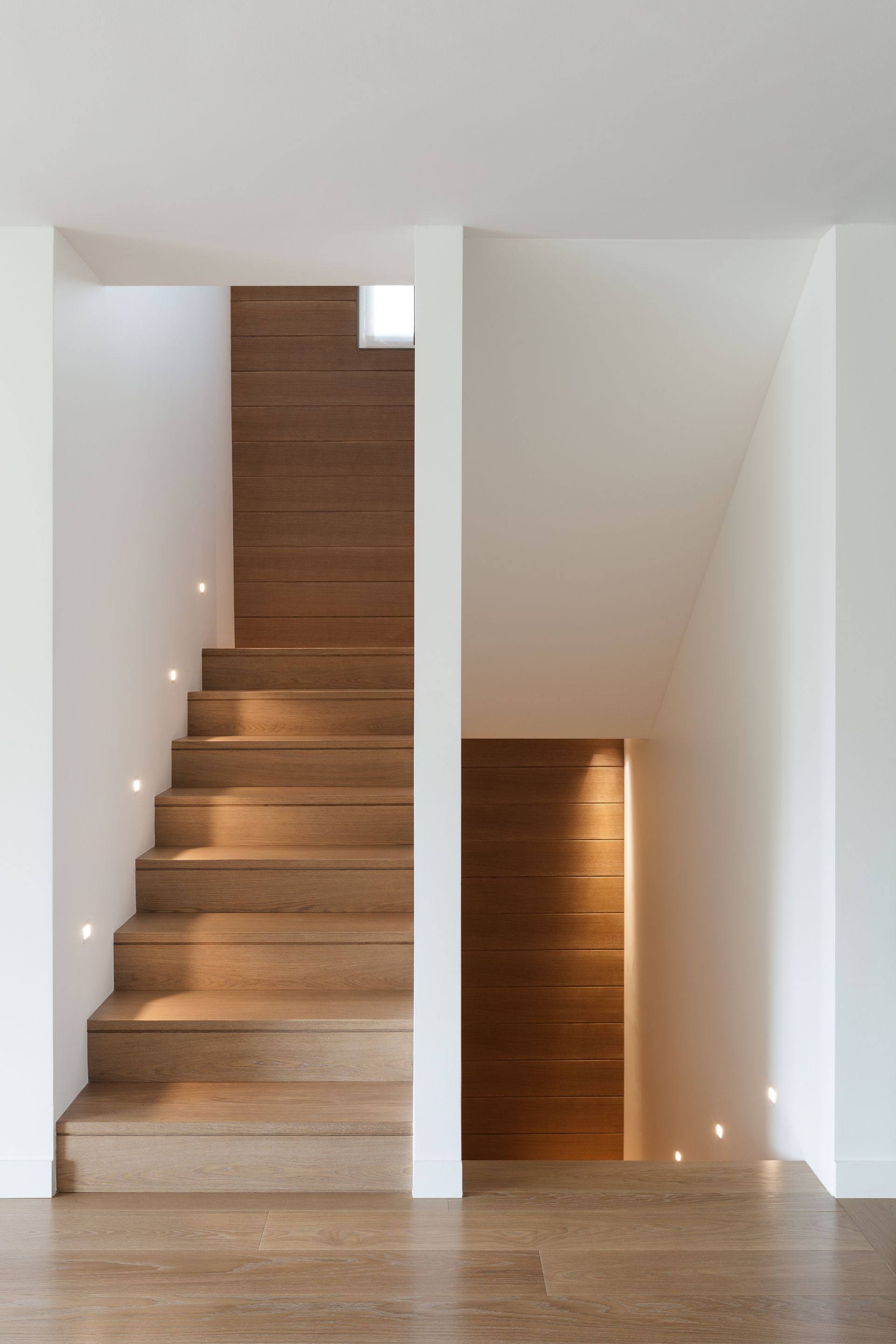 Escalier en bois avec éclairage encastré, sur fond de murs blancs et de lambris bruns.