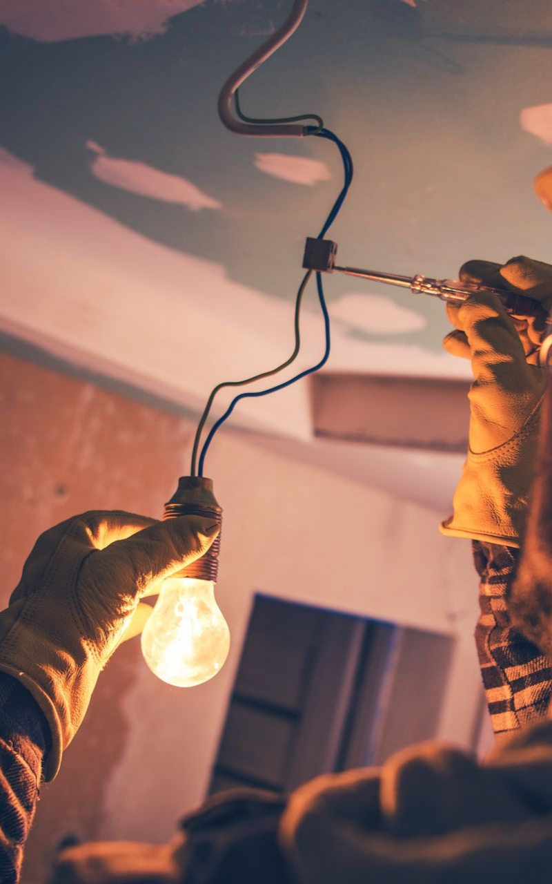 Une personne portant des gants change une ampoule fixée à des fils électriques, travaillant près du plafond.