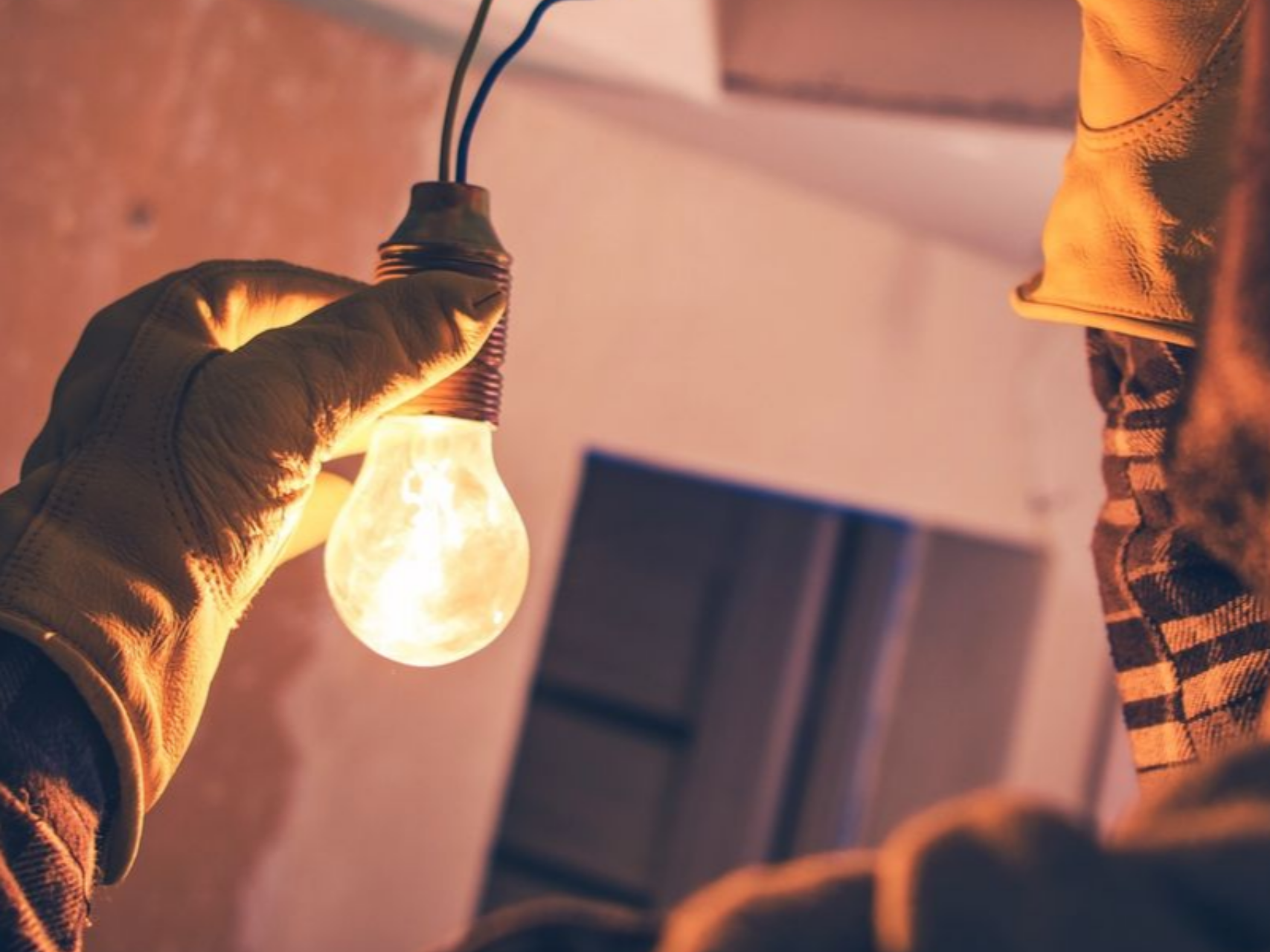Une personne gantée tient une ampoule allumée dont les fils sont dénudés. Intérieur, sur un chantier.