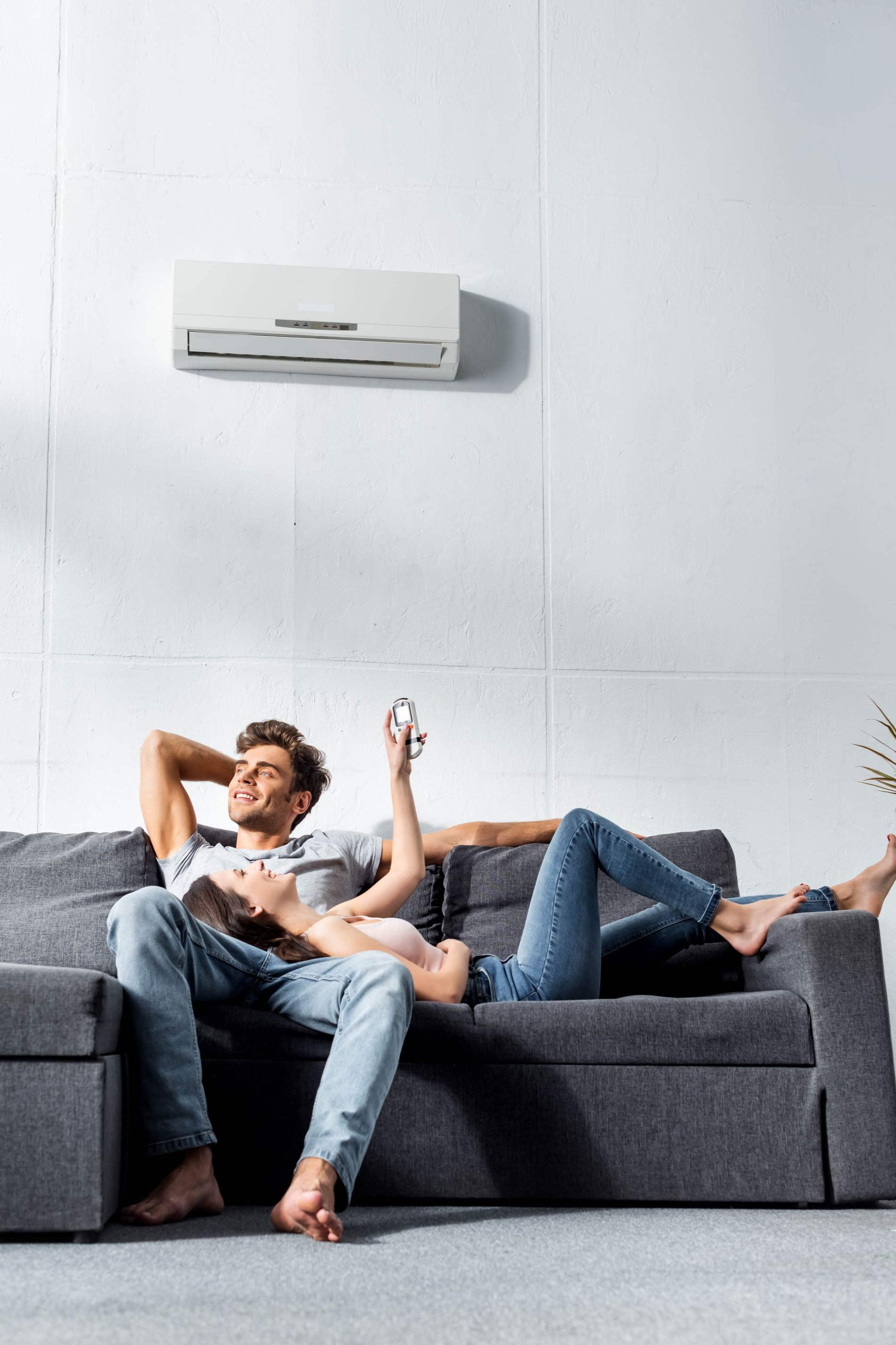 Un couple se détend sur un canapé gris sous un climatiseur. L'un tient une télécommande, tous deux sont pieds nus.