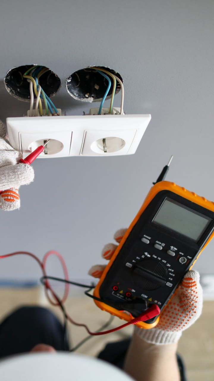 Un électricien utilise un multimètre pour tester une prise électrique blanche dont les fils sont dénudés.