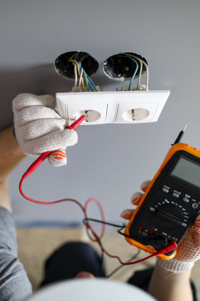 Un électricien utilise un multimètre pour tester une prise électrique blanche dont les fils sont dénudés.