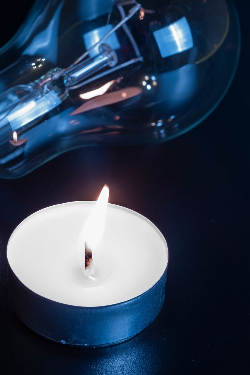Bougie allumée à la flamme vacillante, à côté d'une ampoule éteinte, sur un fond bleu foncé.