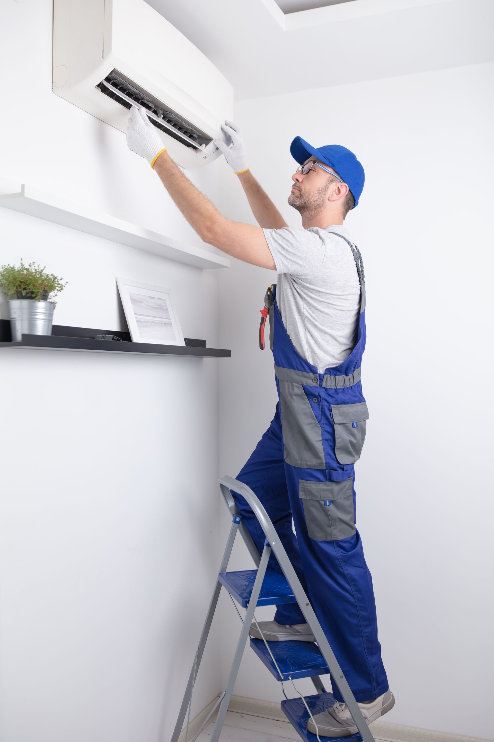 Un homme en salopette bleue, sur une échelle, répare un climatiseur sur un mur blanc.