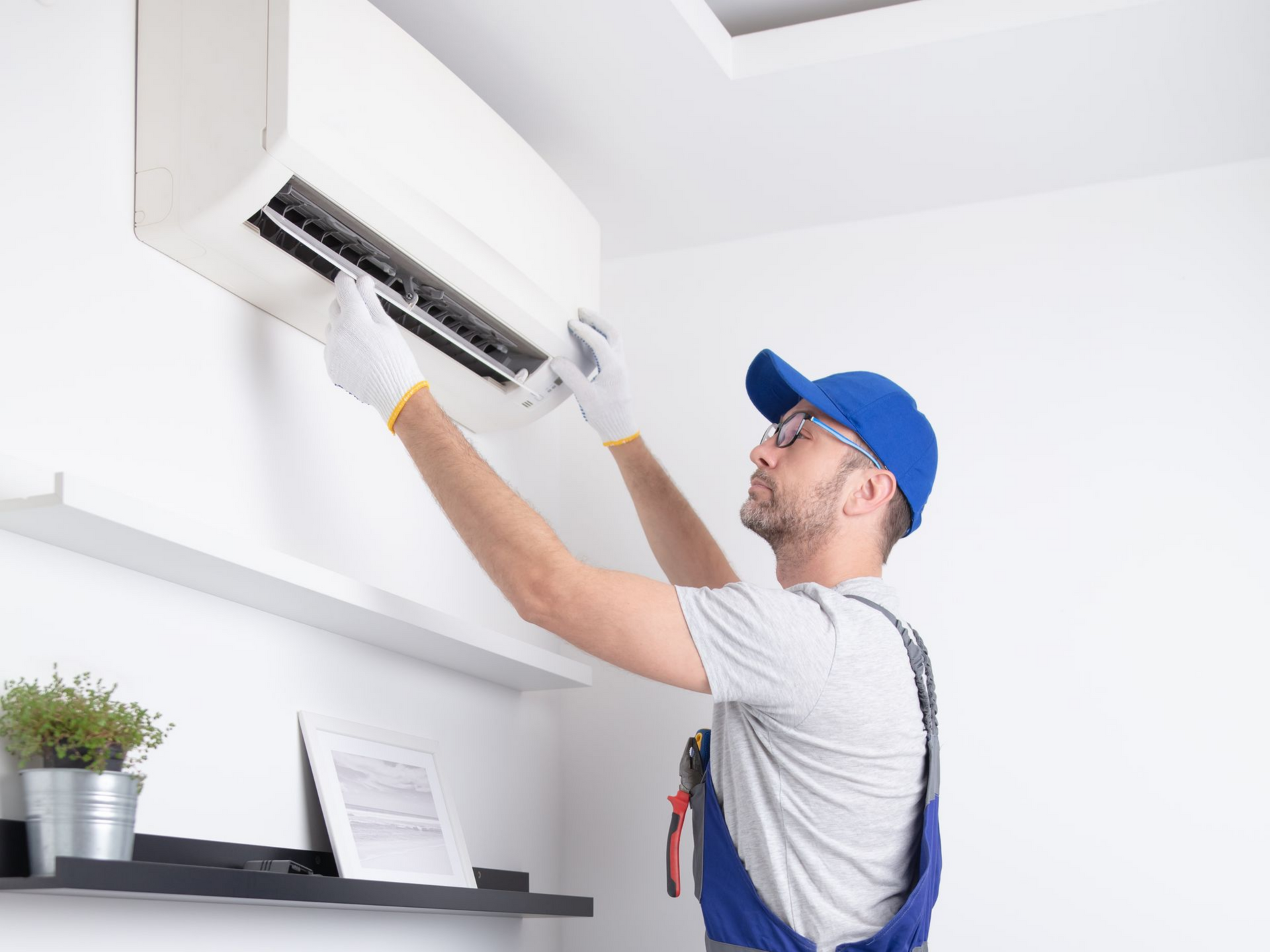 Technicien CVC en uniforme bleu effectuant l'entretien d'un climatiseur mural ; gants blancs, outils.