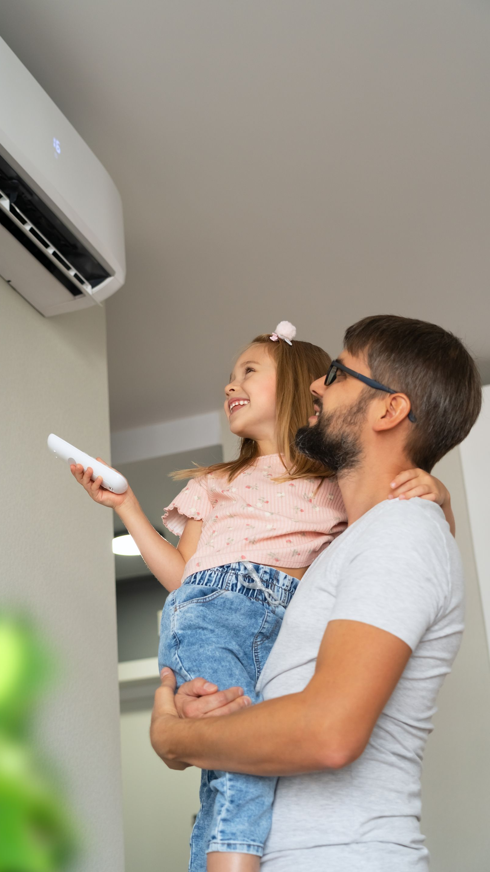 Un homme tenant un enfant dans ses bras, tous deux regardant un climatiseur. L'enfant tient une télécommande et sourit.