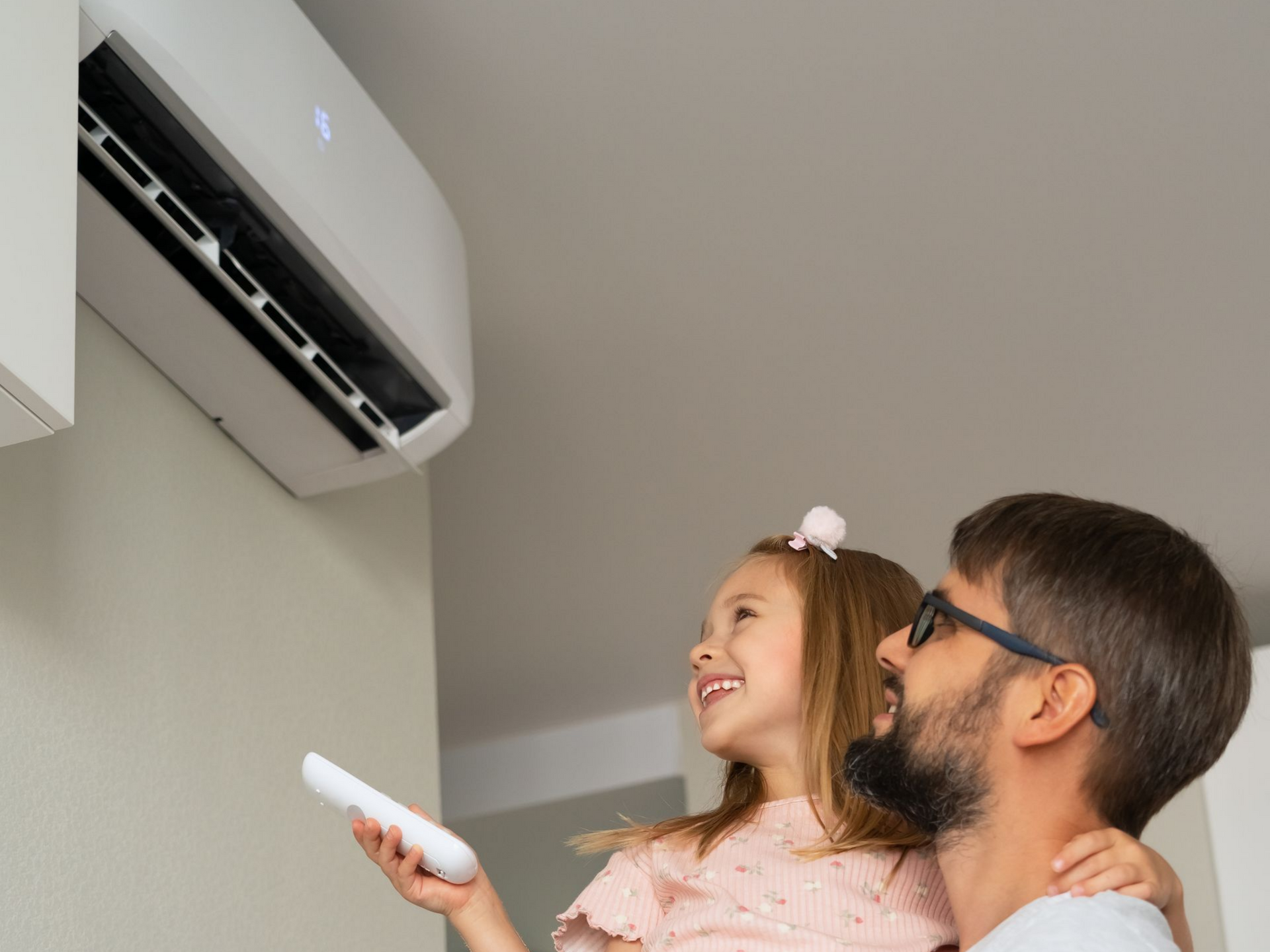 Un père et son enfant regardent un climatiseur mural ; l'enfant tient la télécommande et sourit.