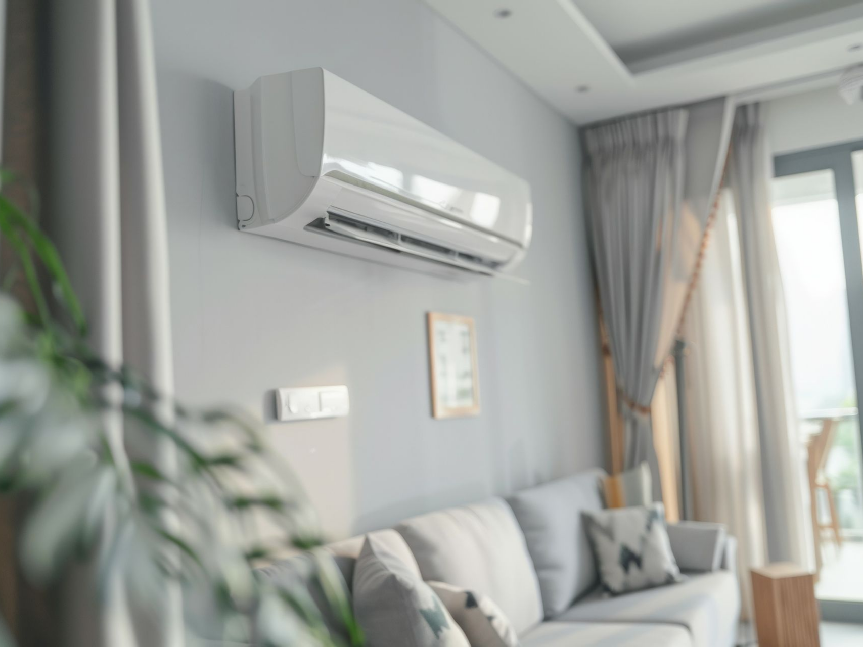 Unité de climatisation blanche fixée sur un mur gris au-dessus d'un canapé. Pièce ensoleillée avec rideaux et plante.