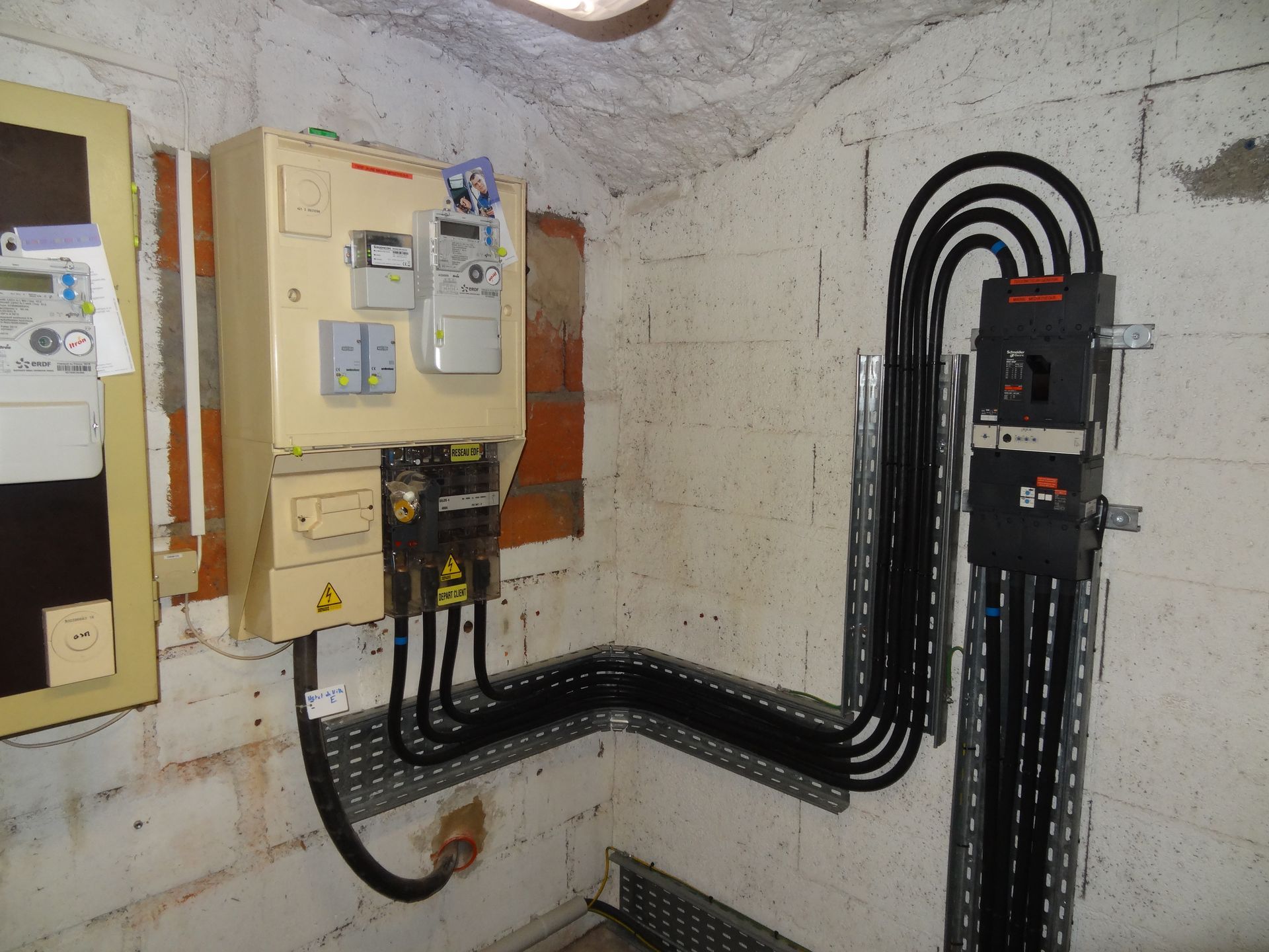 Tableau électrique et compteurs fixés sur un mur de briques. Des câbles noirs courent le long de chemins de câbles métalliques.